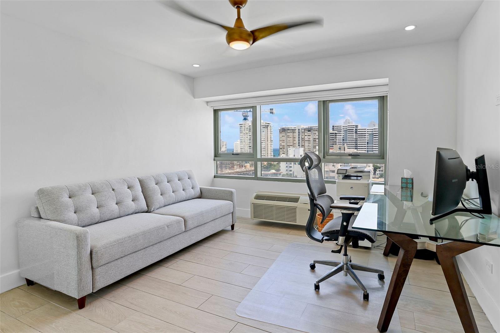 Furnished apartment office with a gray sofa, glass desk, and city skyline views.