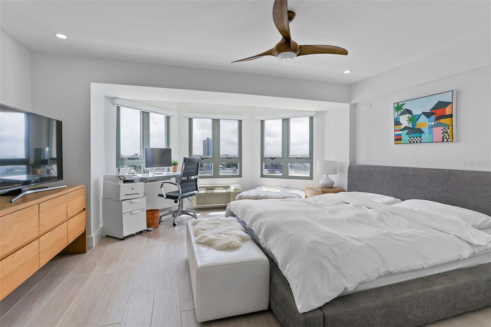 Furnished bedroom with a gray bed, white bedding, and wide windows overlooking the city.