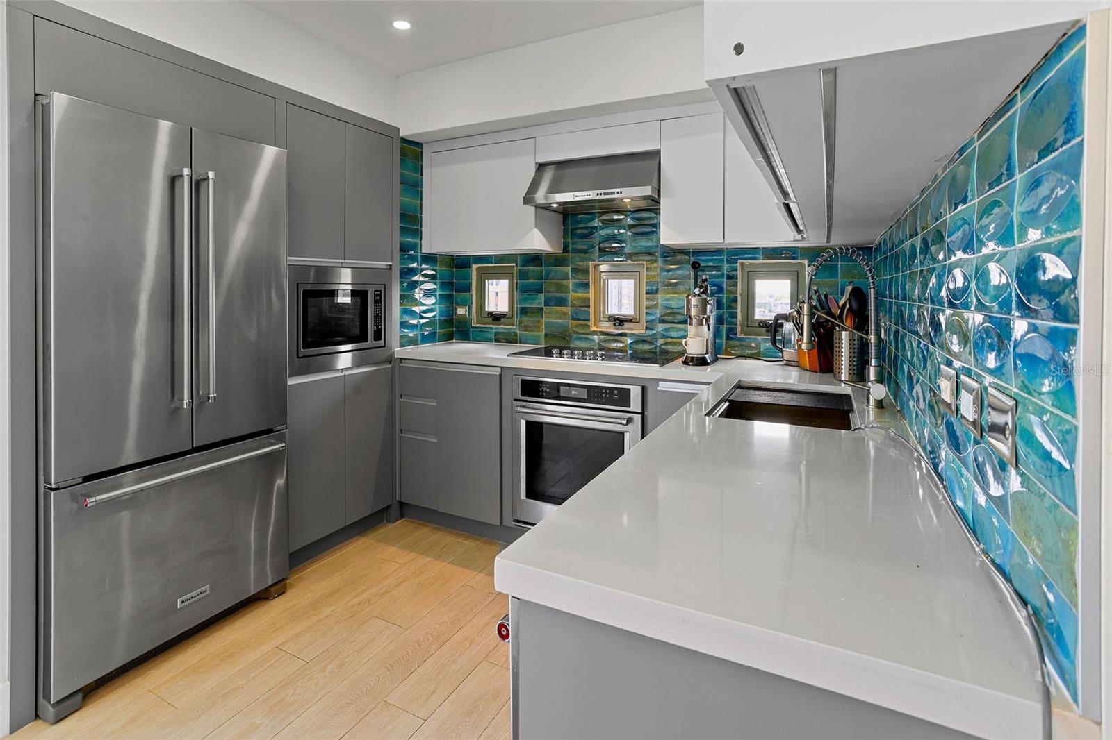 Modern kitchen with stainless steel appliances, white counters, and a glossy blue-green tile backsplash.