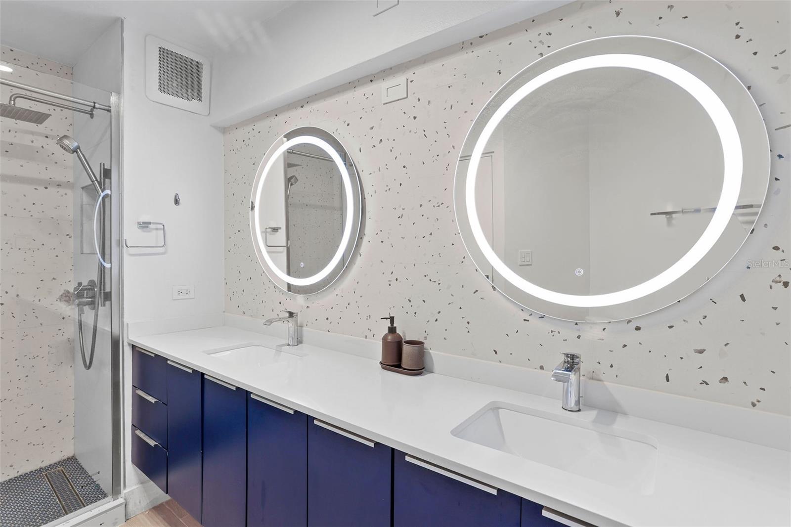 Modern bathroom with double sinks, illuminated round mirrors, dark blue cabinets, and a glass-enclosed shower.