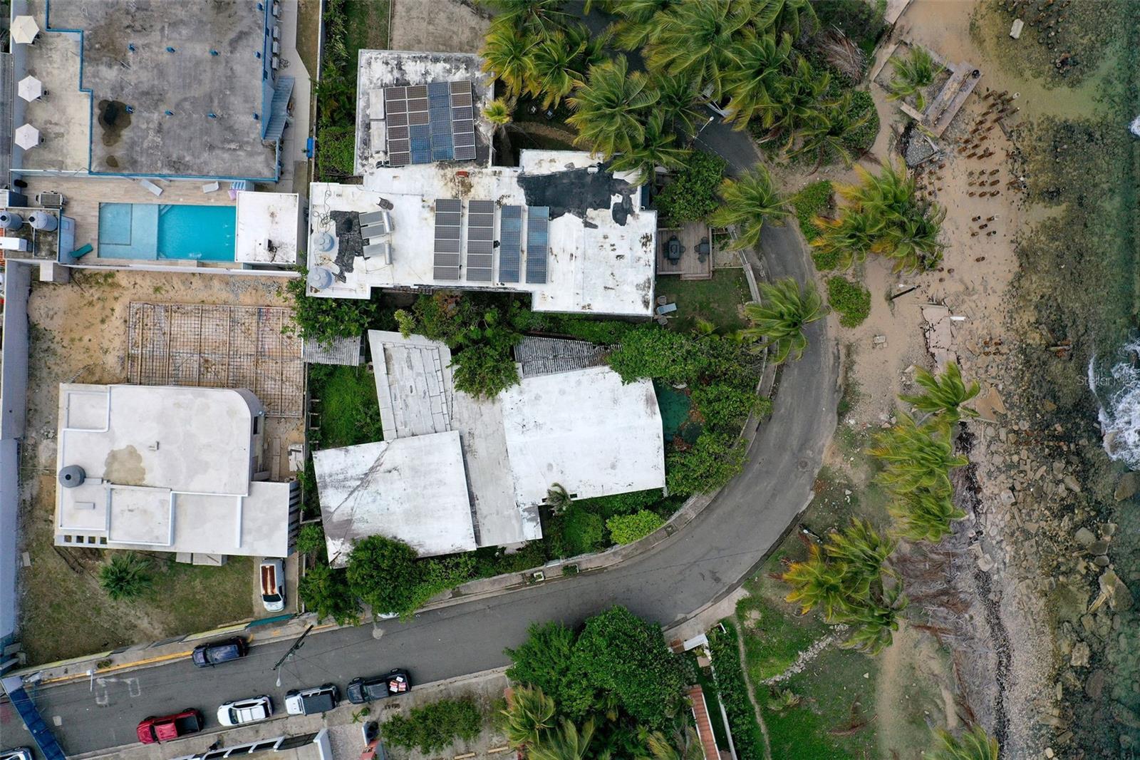 Aerial view of a coastal property with rooftop solar panels beside a curving road and rocky shoreline.