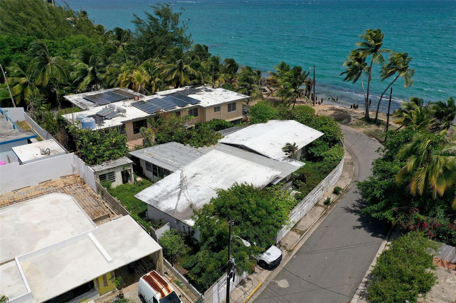 Coastal homes beside turquoise water, with solar panels on a flat-roofed house and palms along the shore.