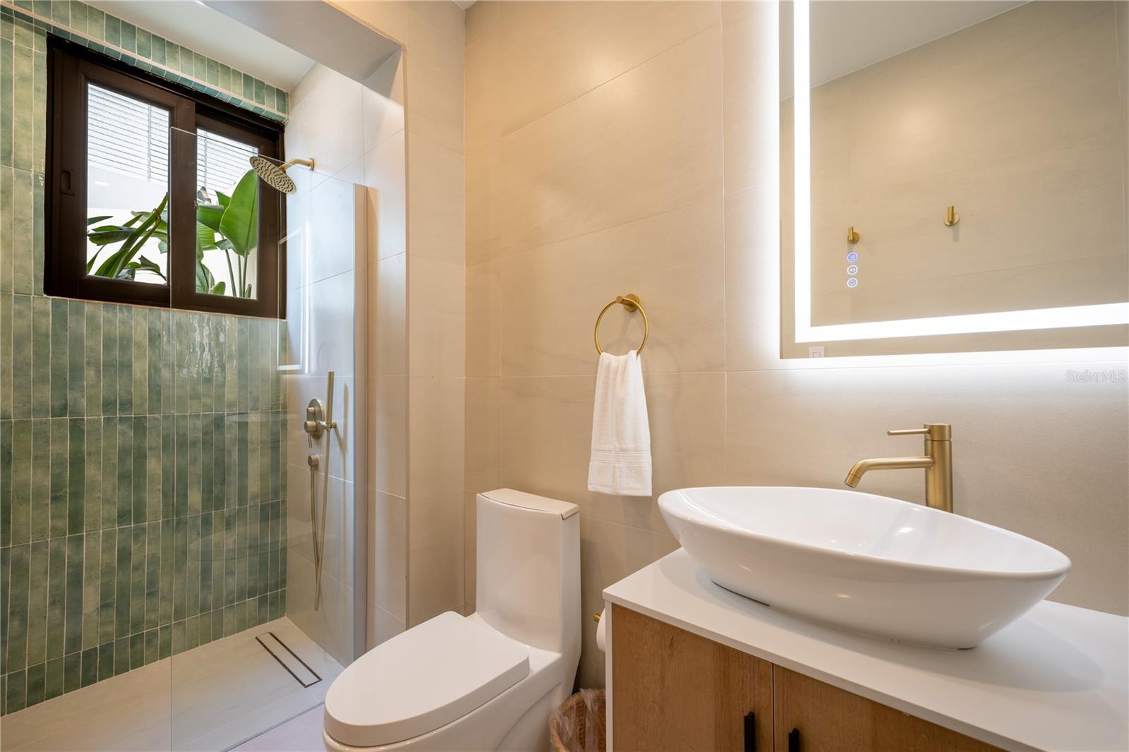 Modern bathroom with a green-tiled shower, vessel sink, brass fixtures, and illuminated mirror.
