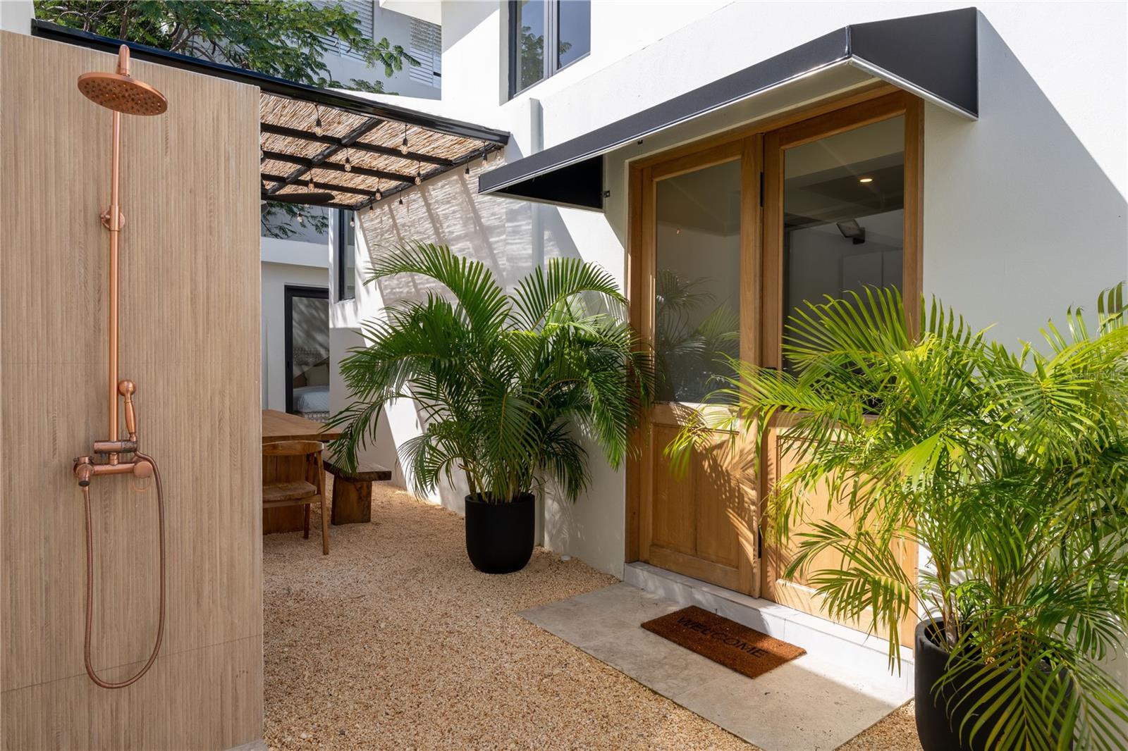 Outdoor entry courtyard with a copper shower, tropical palms, and wood-framed glass doors.