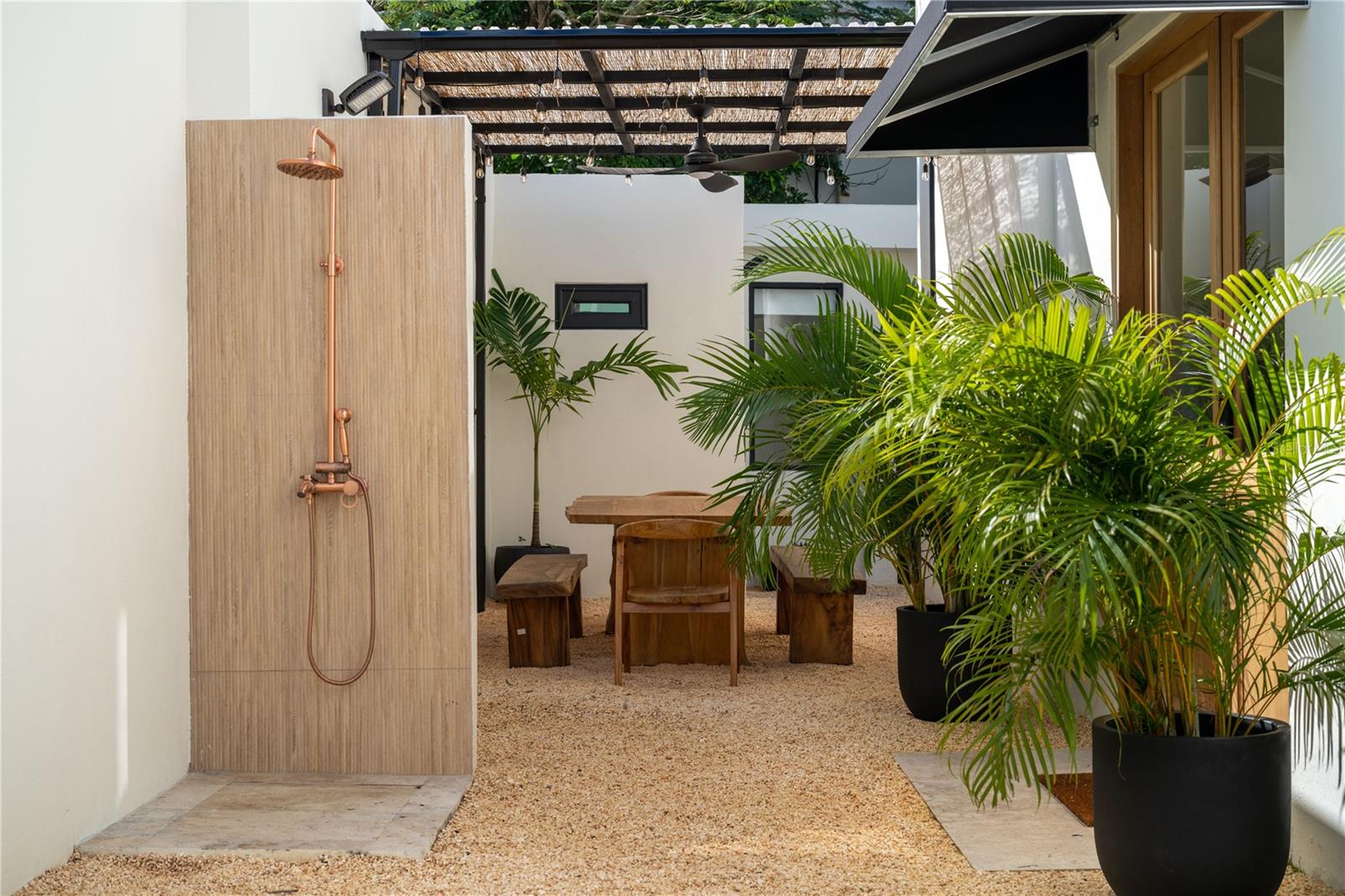 Outdoor patio with a copper shower, wooden seating, potted palms, and a slatted pergola.