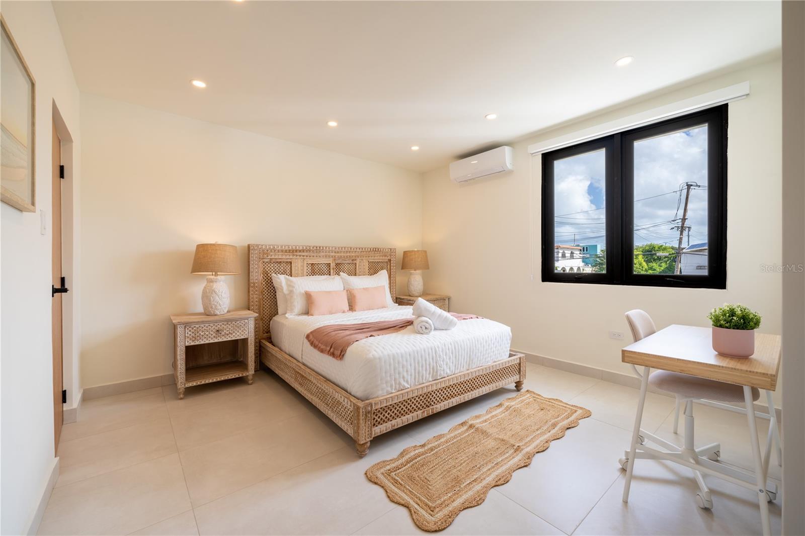 Furnished bedroom with a woven bed, blush accents, and a desk beside a large window.