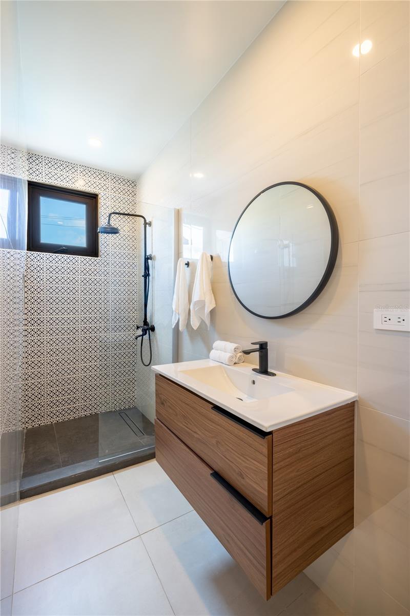 Modern bathroom with geometric-tile walk-in shower, wood vanity, and large round mirror.