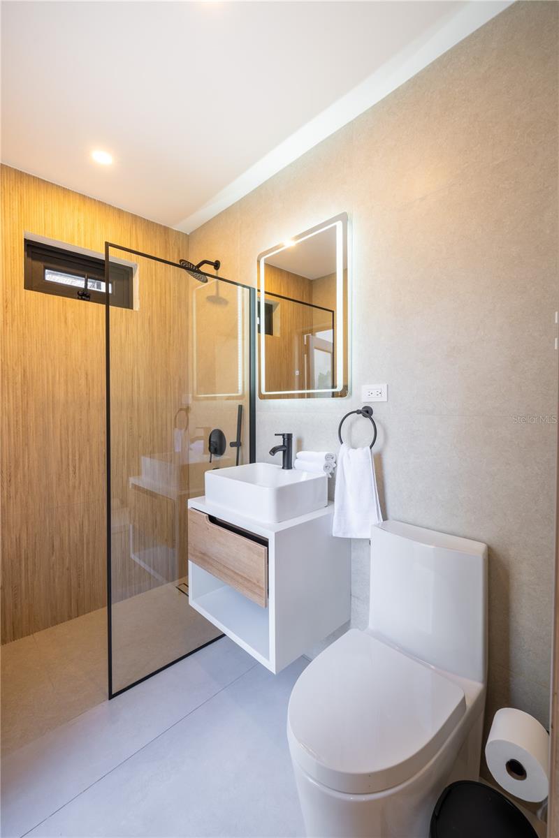 Modern bathroom with a glass shower, wood-look walls, floating vanity, illuminated mirror, and white toilet.