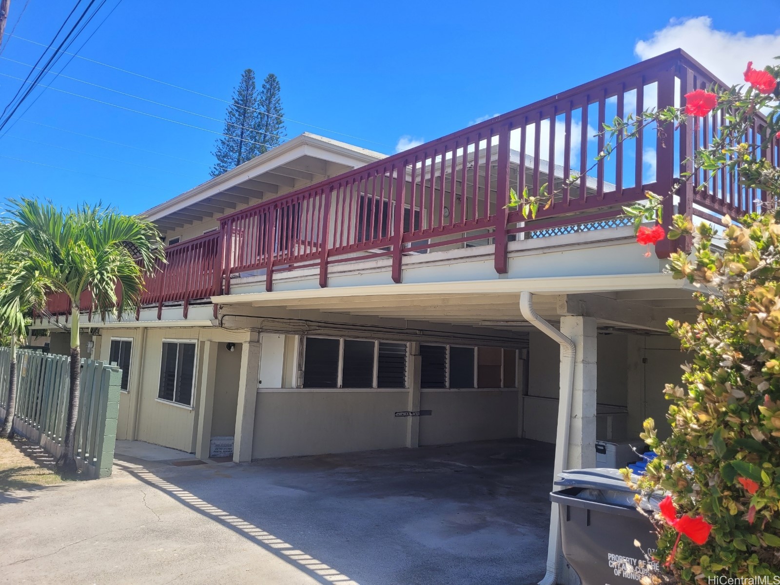 Raised home with a long burgundy deck above a shaded carport, framed by a palm and red hibiscus.