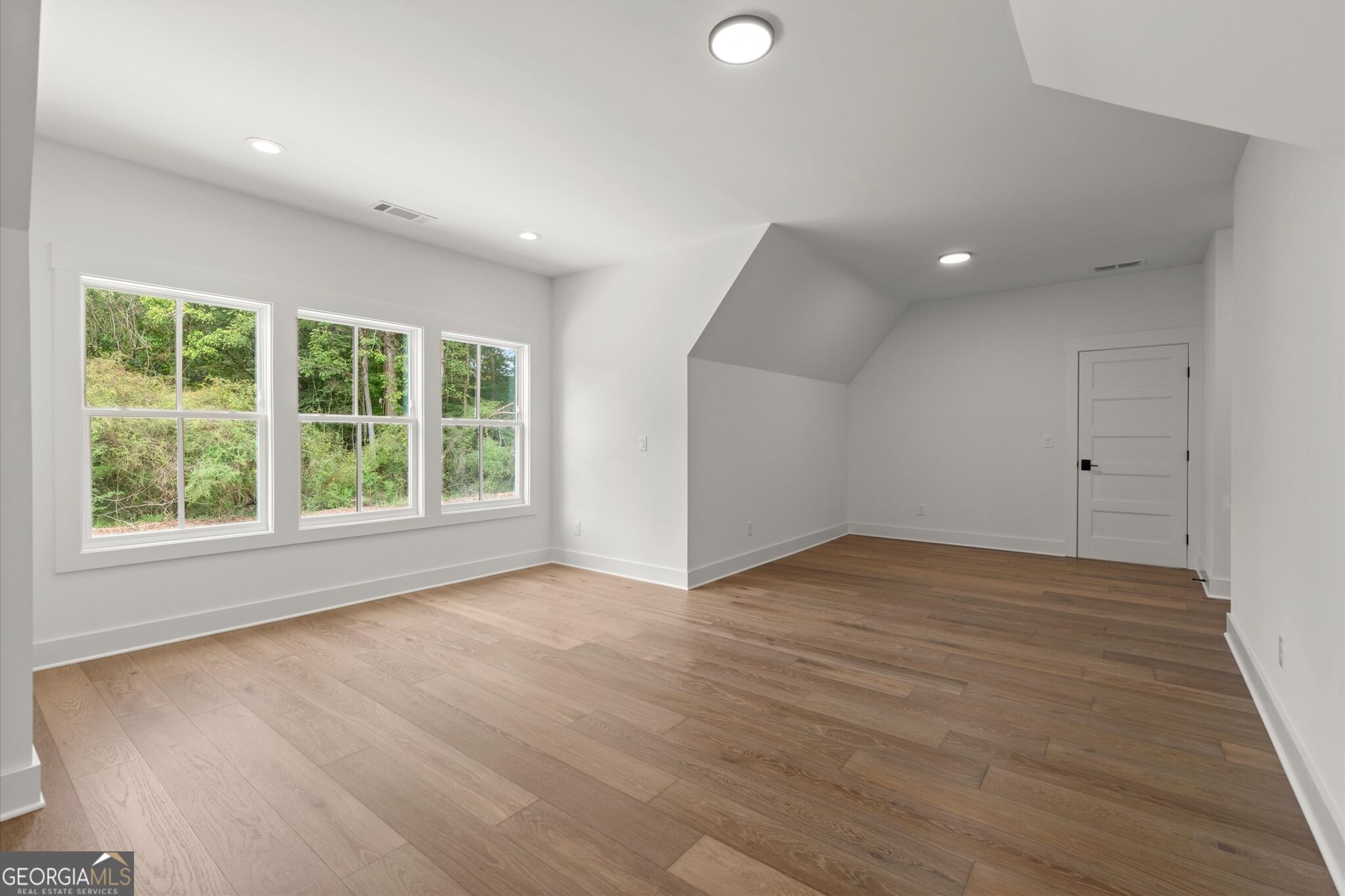 Empty room with wood flooring, three windows overlooking trees, and a sloped ceiling.