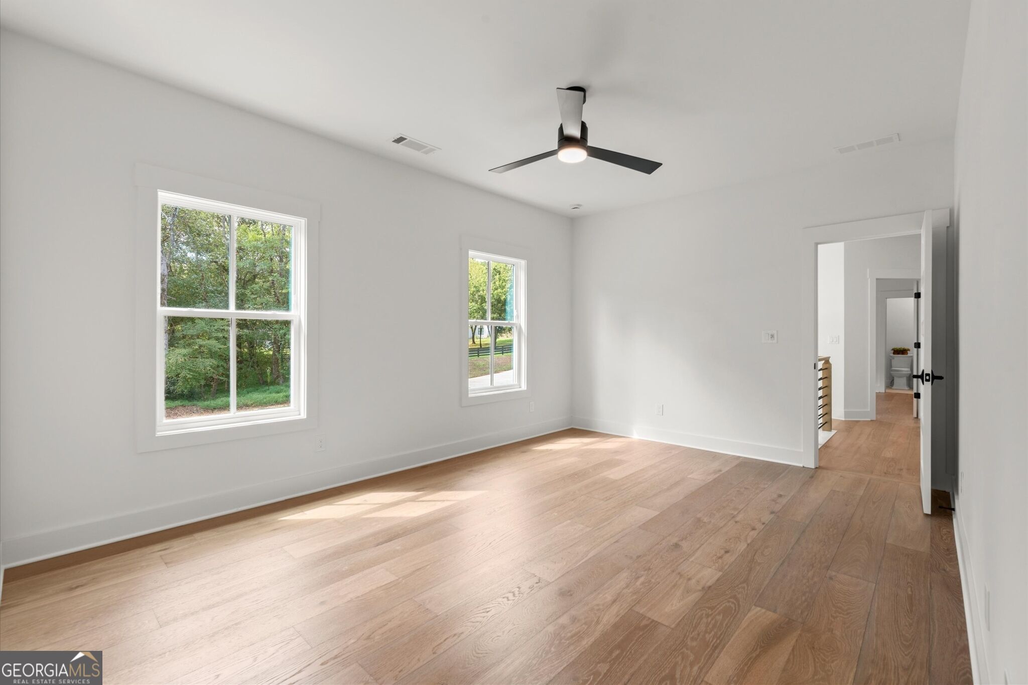 Vacant room with light wood floors, two windows, and a black ceiling fan.
