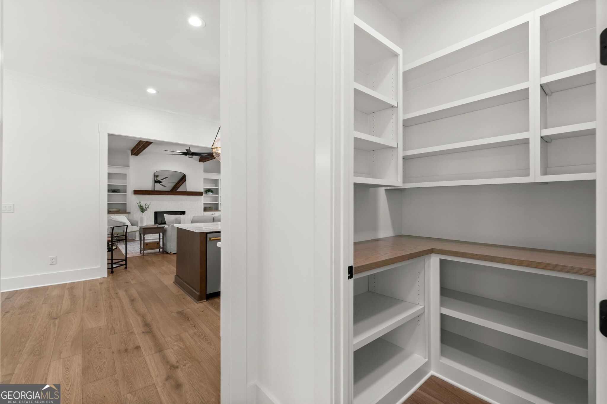 Walk-in pantry with white built-in shelves and a wood countertop, opening toward the kitchen and living area.