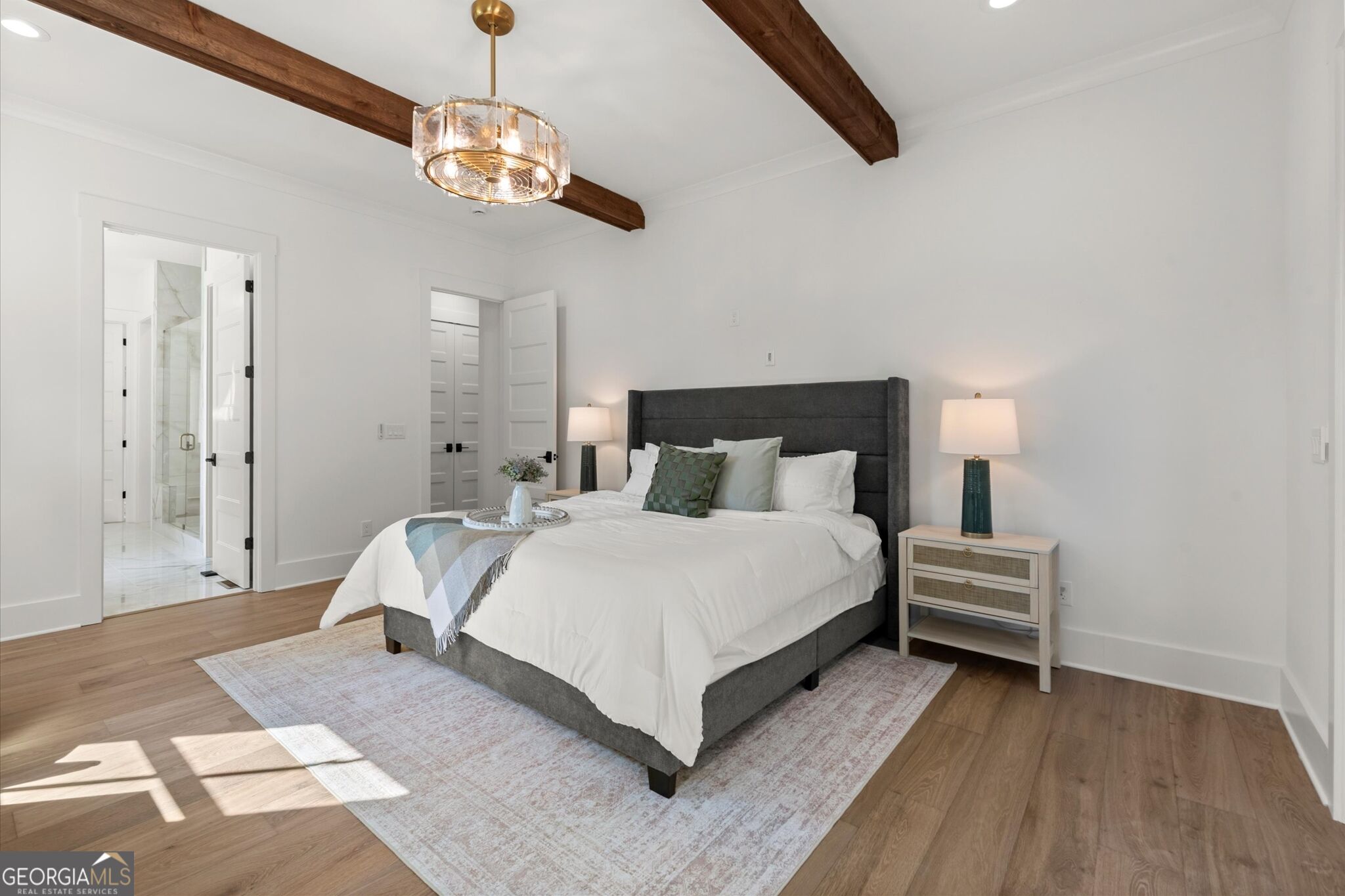 Staged bedroom with a gray upholstered bed, exposed wood ceiling beams, and an open bathroom doorway.