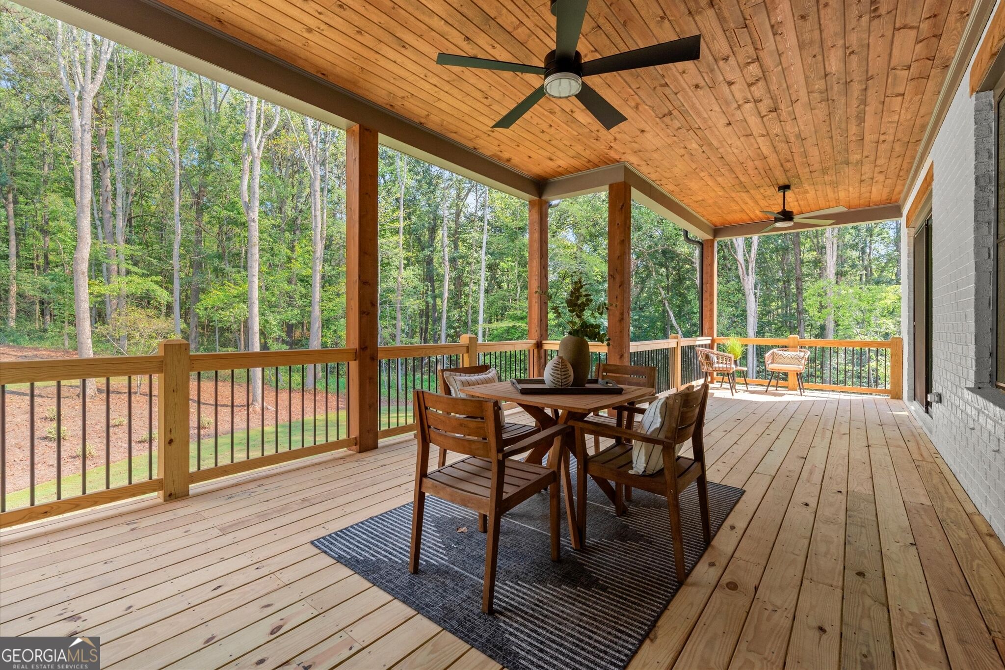 Covered wood deck with dining set and ceiling fans, overlooking a wooded yard.