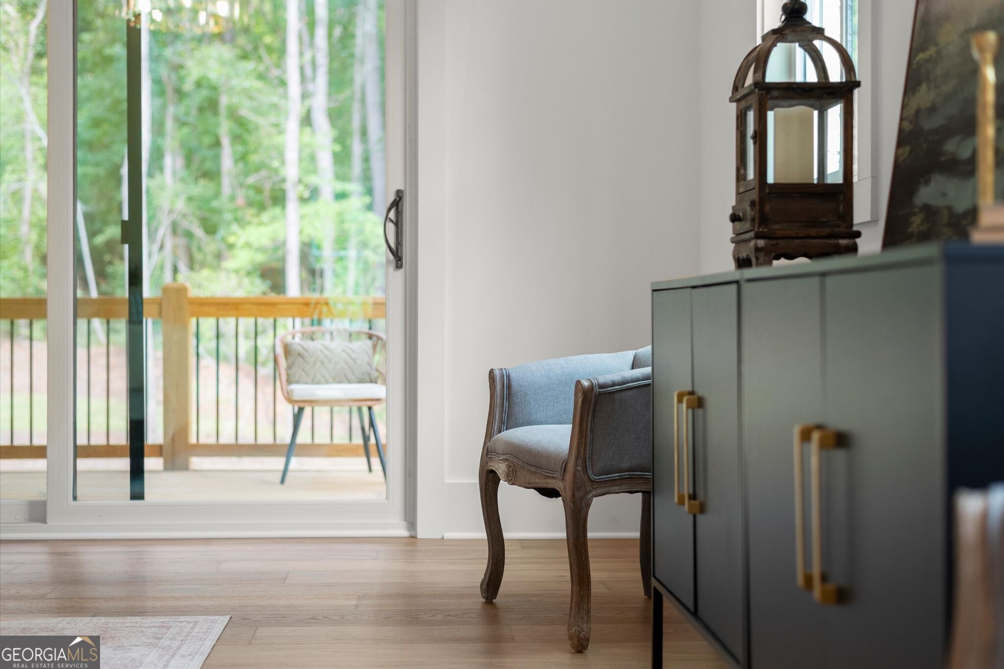 Furnished room with a gray chair and dark cabinet beside sliding glass doors opening to a wooded deck.