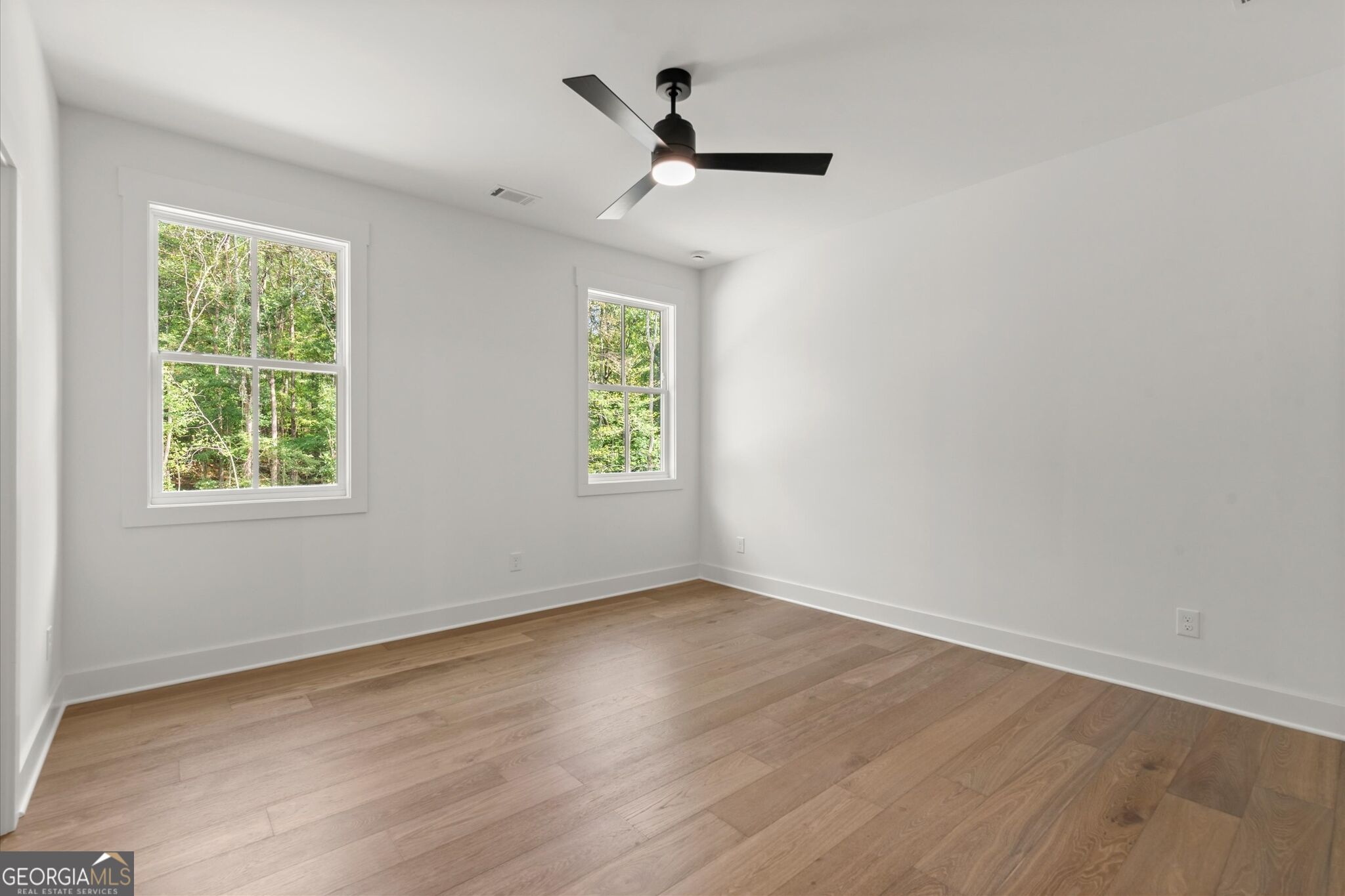 Empty room with light wood floors, two tree-view windows, and a black ceiling fan.