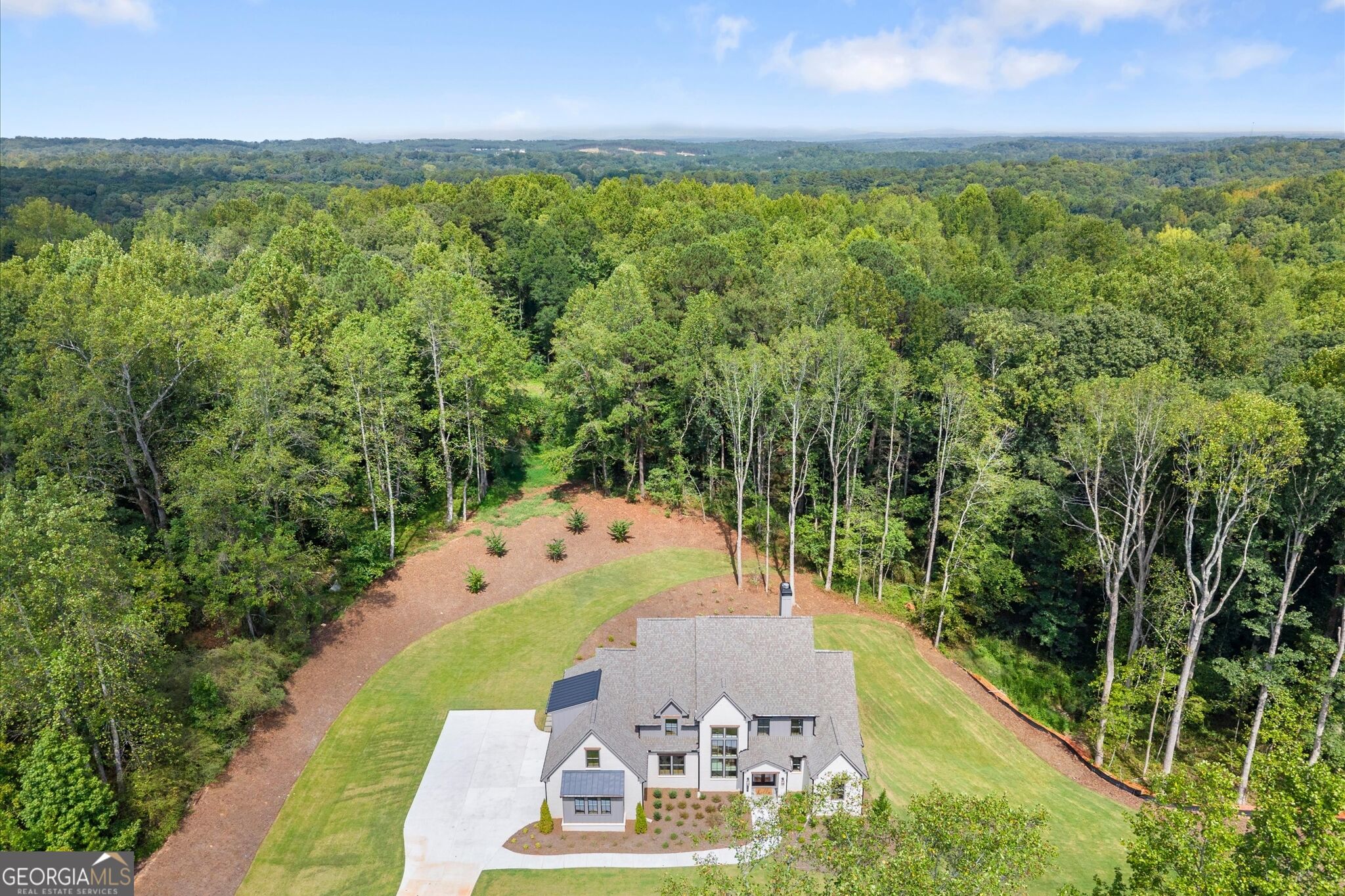 Aerial view of a home surrounded by woodland, with a broad lawn, driveway, and landscaped grounds.