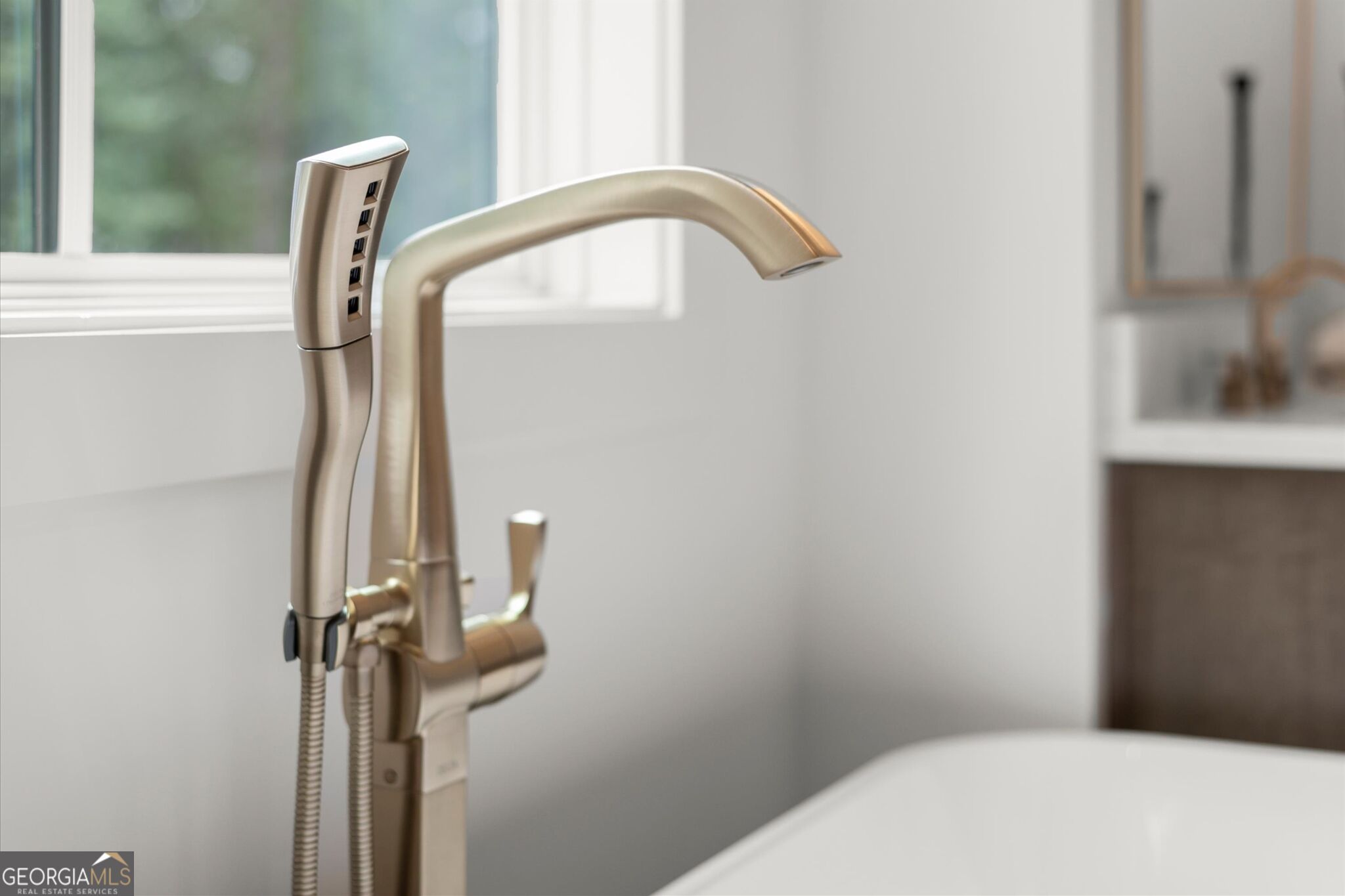 Brushed metal bathtub faucet with handheld sprayer beside a white tub and window.