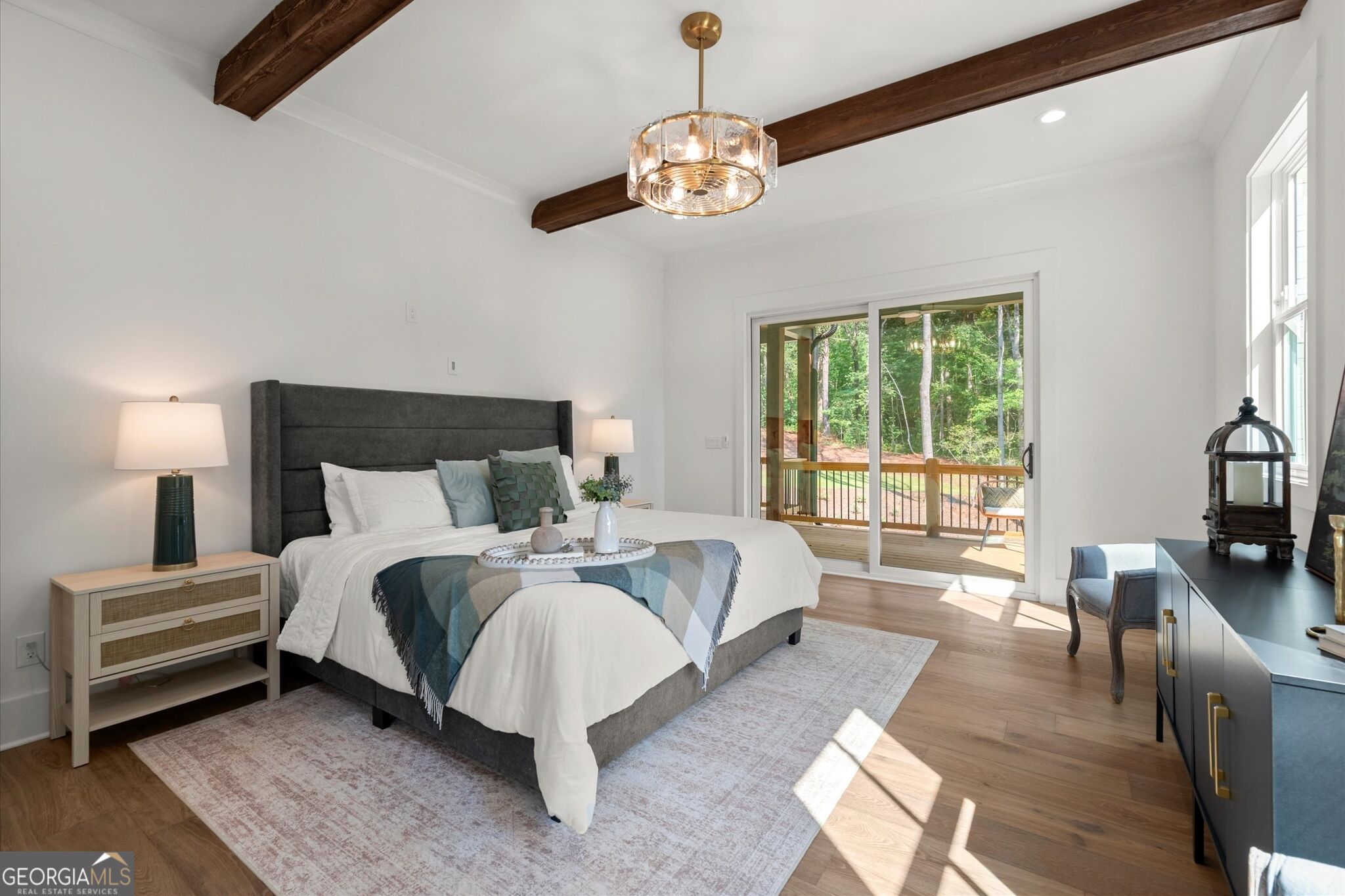 Staged bedroom with dark ceiling beams, a gray bed, and sliding doors opening onto a wooded deck.