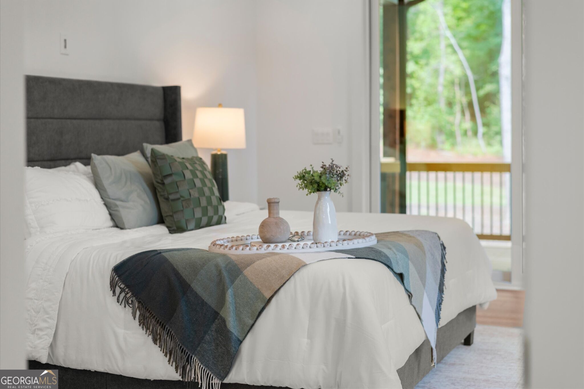 Staged bedroom with a white bed, gray headboard, green pillows, and a view of the deck.