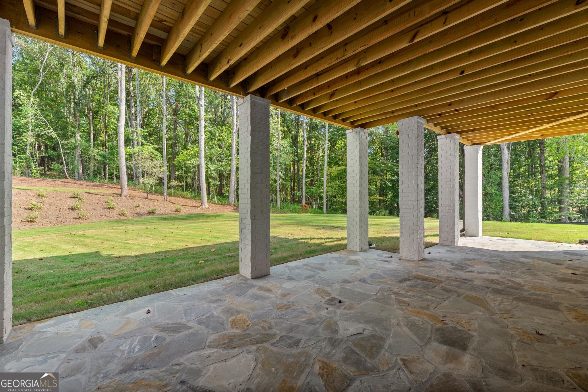 Covered stone patio with a timber ceiling and brick columns overlooking a lawn and woods.