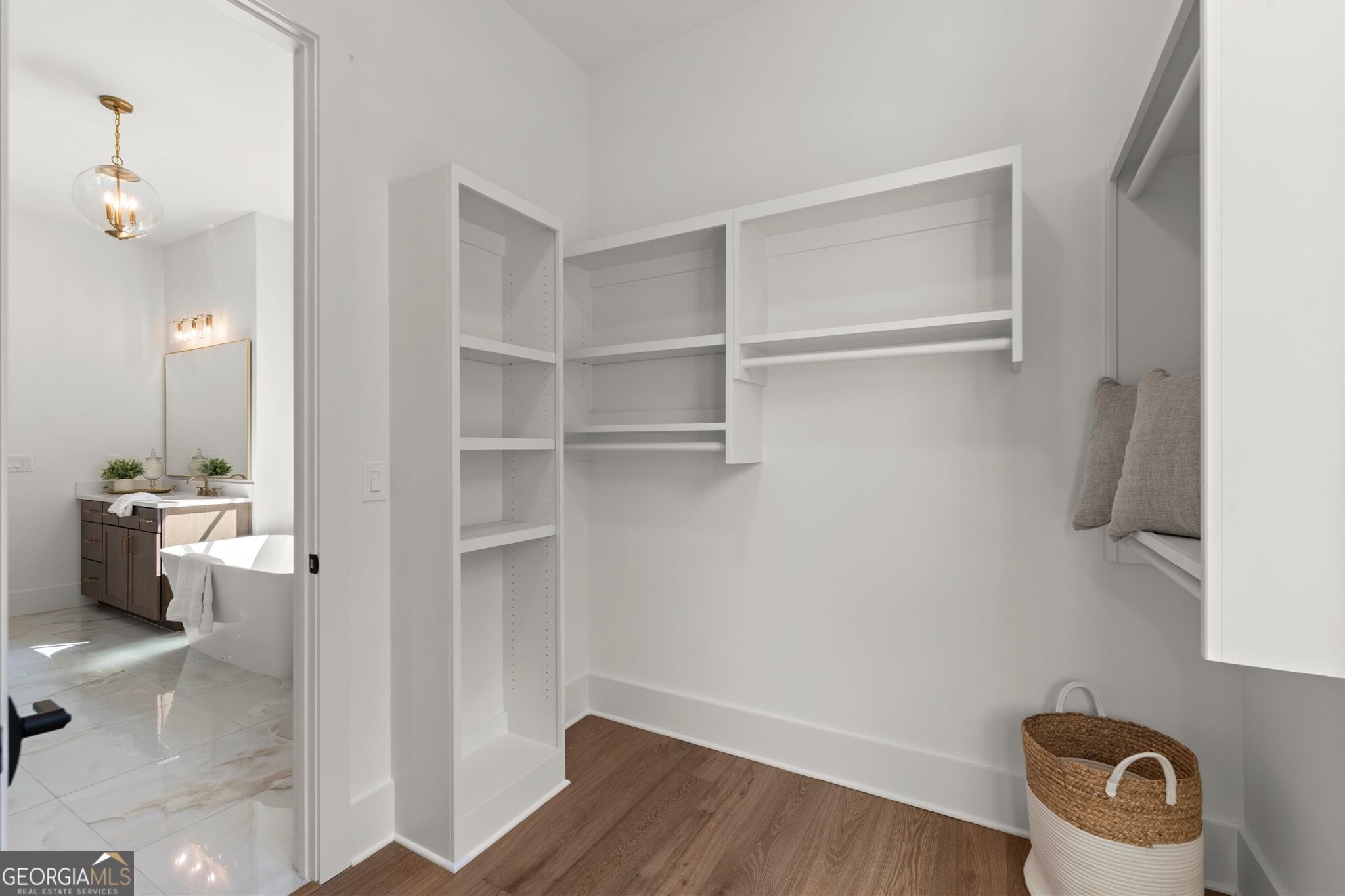 Walk-in closet with white built-in shelves, a cushioned bench, and wood flooring beside a bathroom.