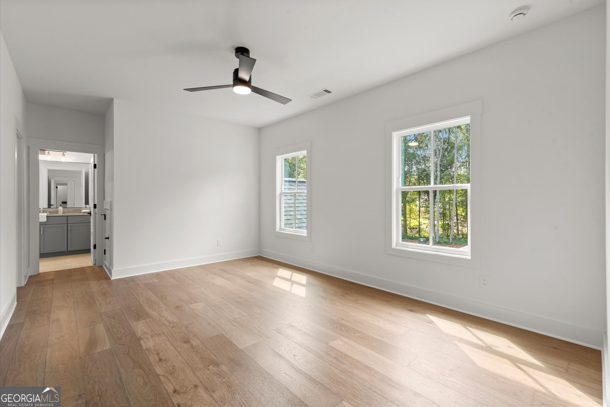 Empty bedroom with wood floors, two windows overlooking trees, and an adjoining bathroom.