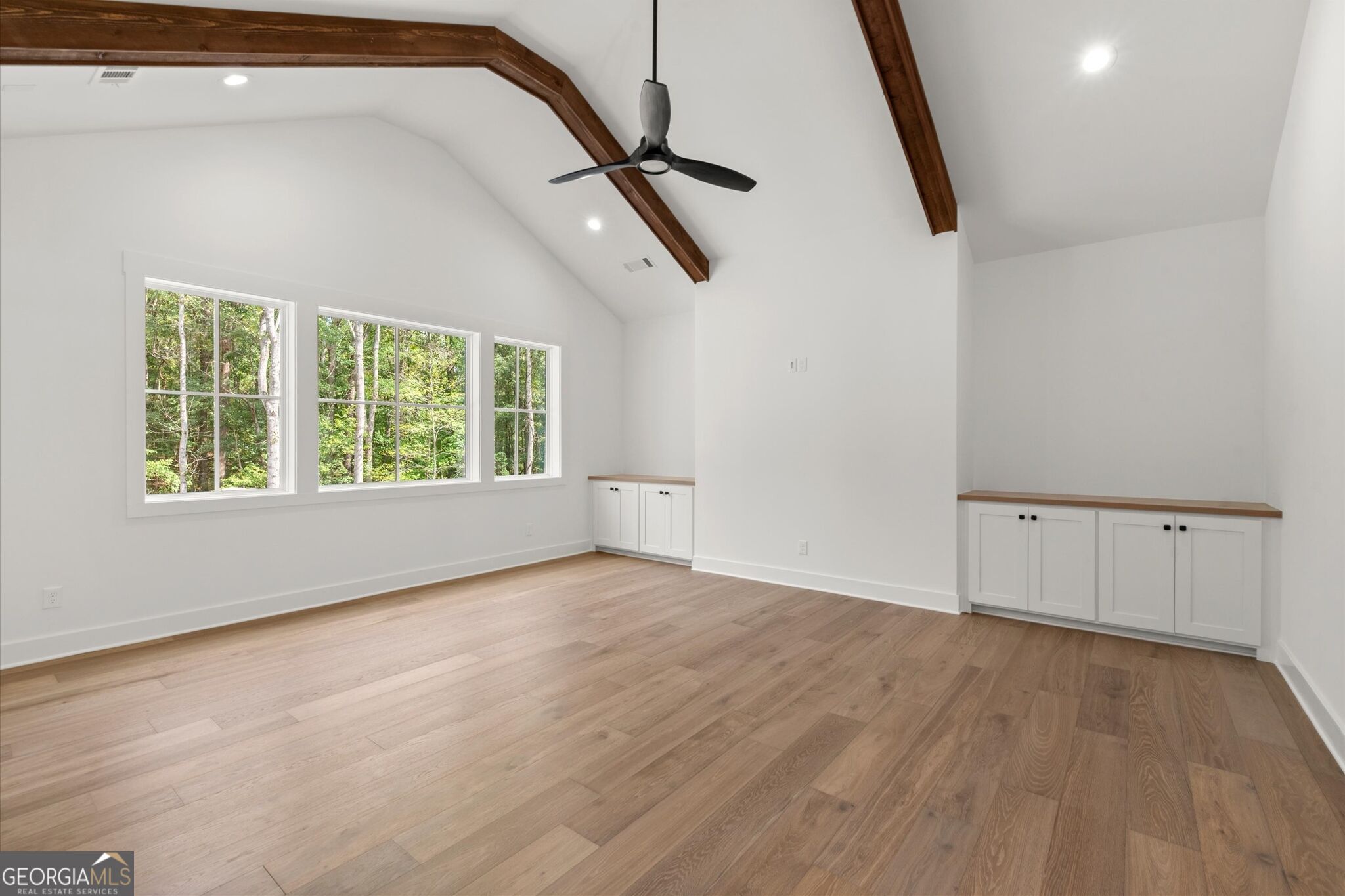 Vacant room with vaulted ceiling, exposed wood beams, built-in cabinets, and windows overlooking trees.