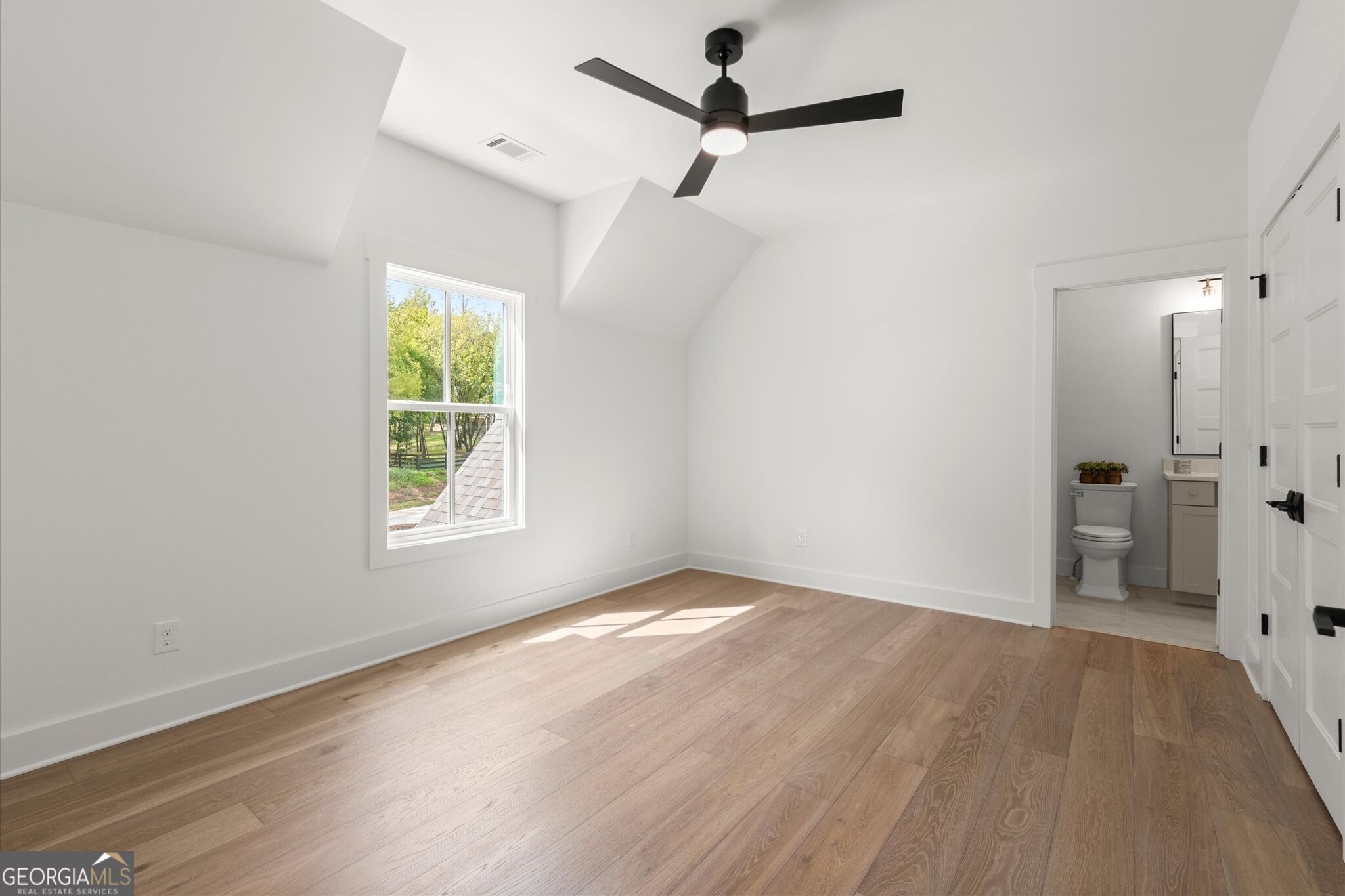 Vacant bedroom with vaulted ceiling, black ceiling fan, wood floors, and an adjoining bathroom.