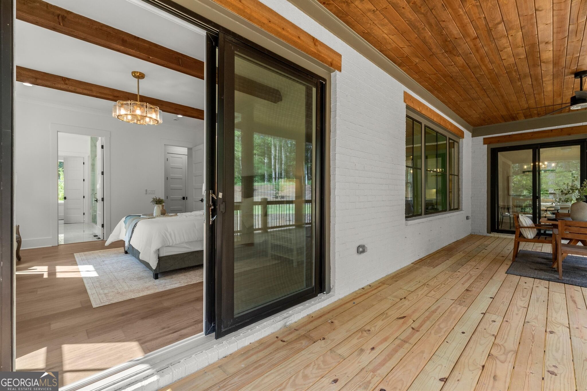 Covered wooden deck beside a bedroom, with a plank ceiling and black-framed sliding glass doors.