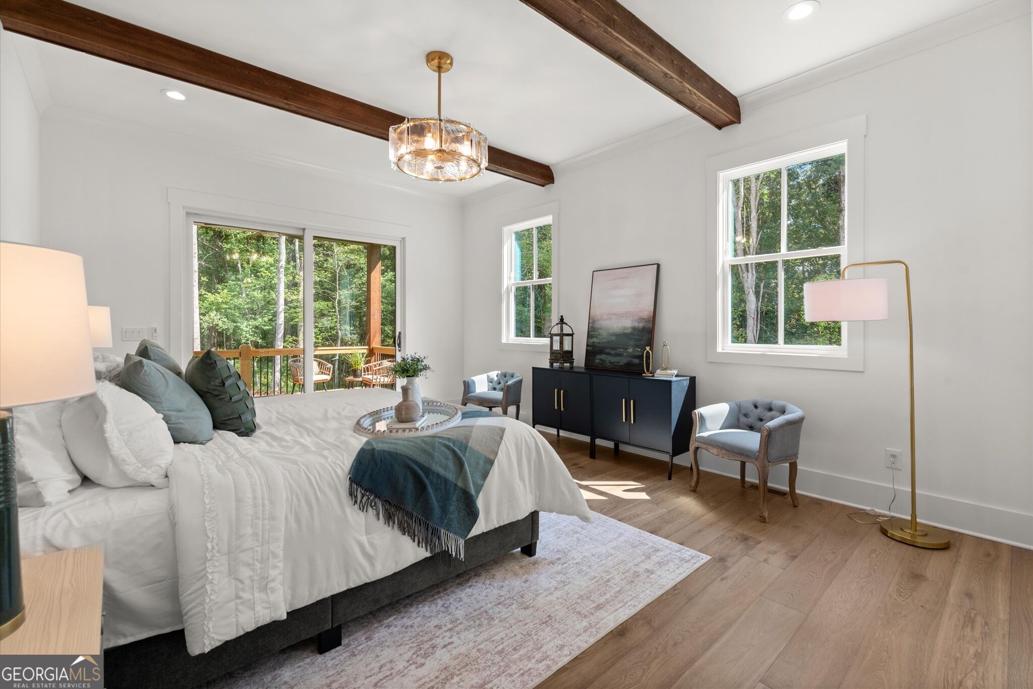 Staged bedroom with exposed beams, a large bed, and sliding doors to a wooded deck.
