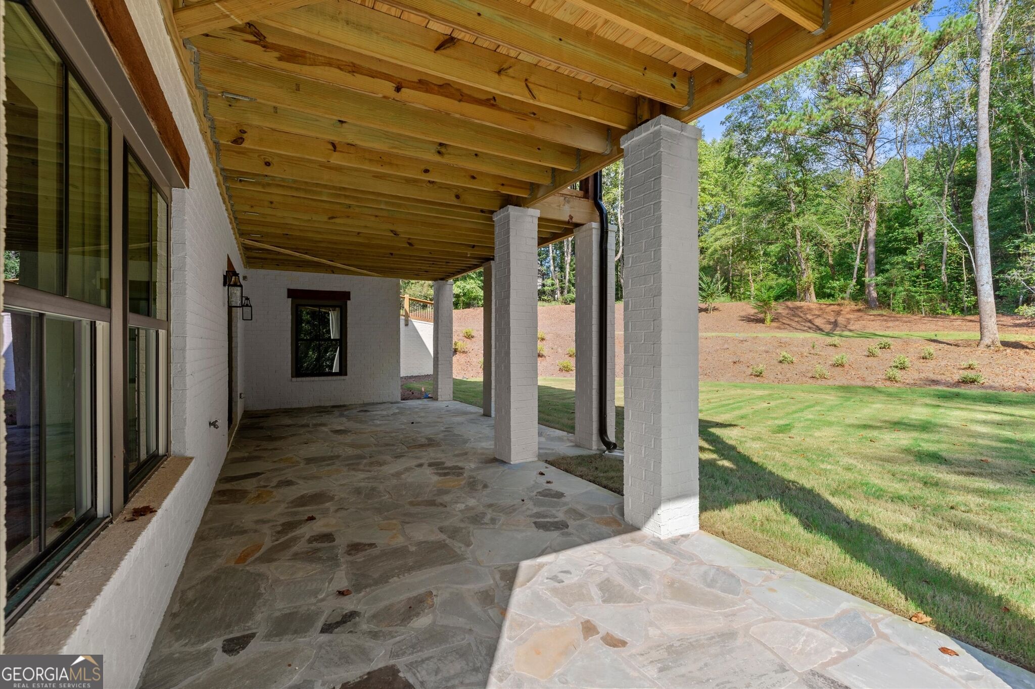 Covered patio with a stone floor, exposed wood ceiling, and white brick columns beside a grassy yard.