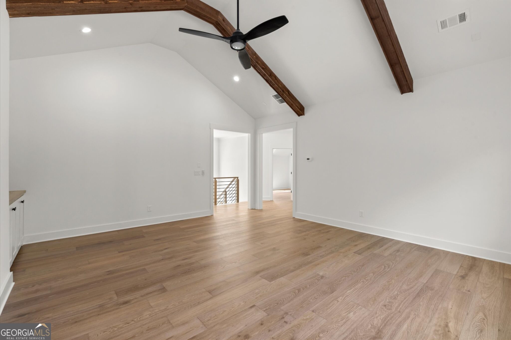 Empty room with vaulted ceiling, exposed wood beams, black fan, and light wood floors.