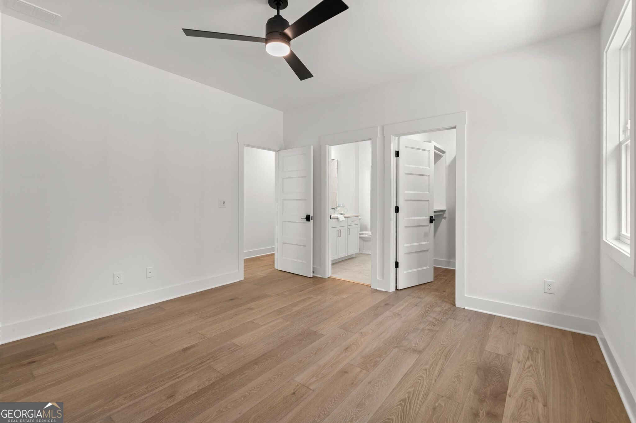 Empty bedroom with wood floors, a black ceiling fan, and open doors to a bathroom and adjoining space.