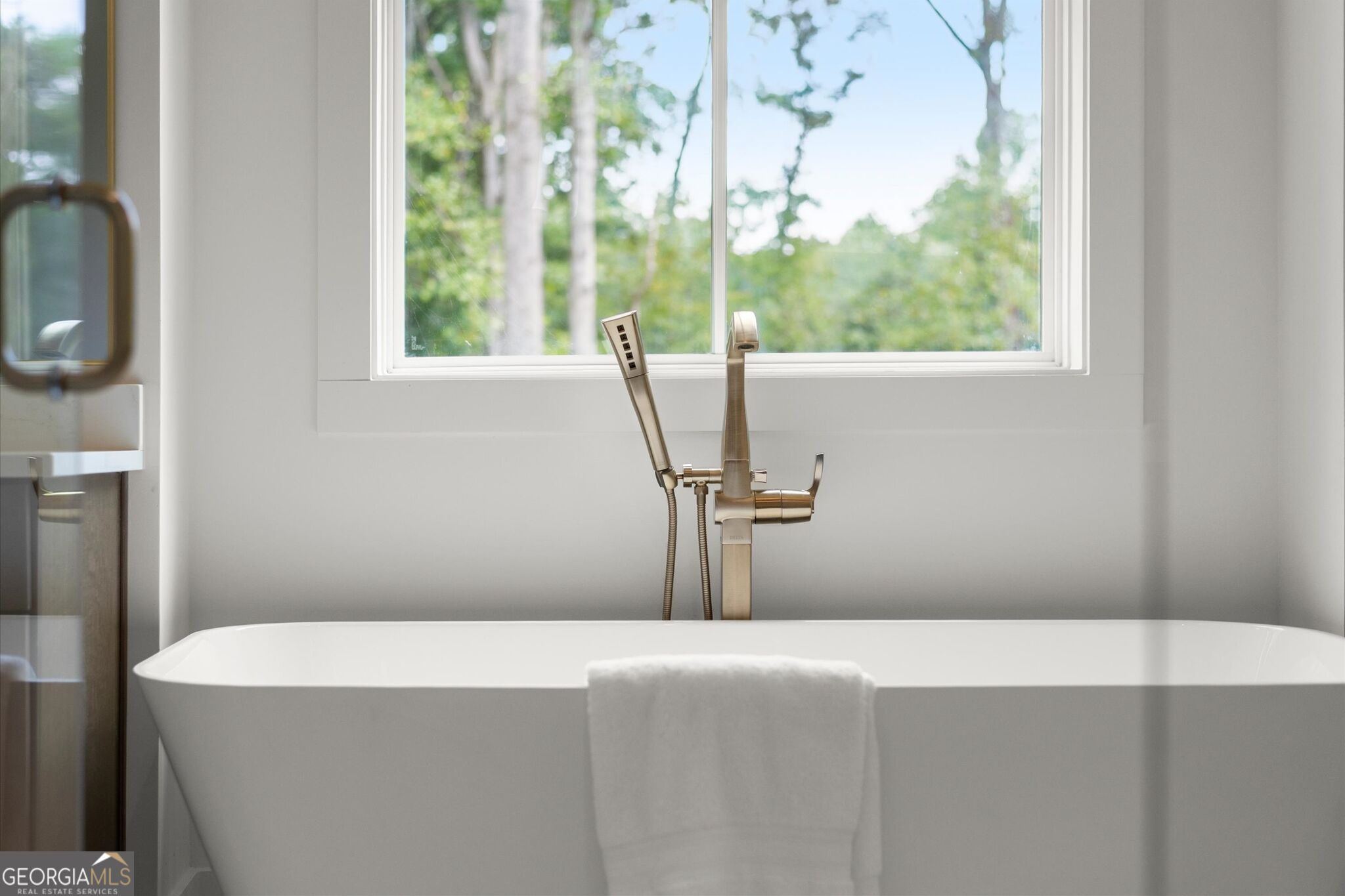 Freestanding bathtub with brass fixtures beneath a large window overlooking trees.