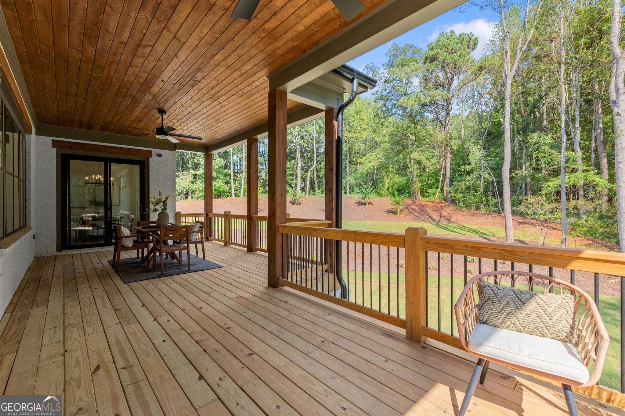 Covered wooden deck with dining furniture and a wicker chair overlooking a wooded backyard.