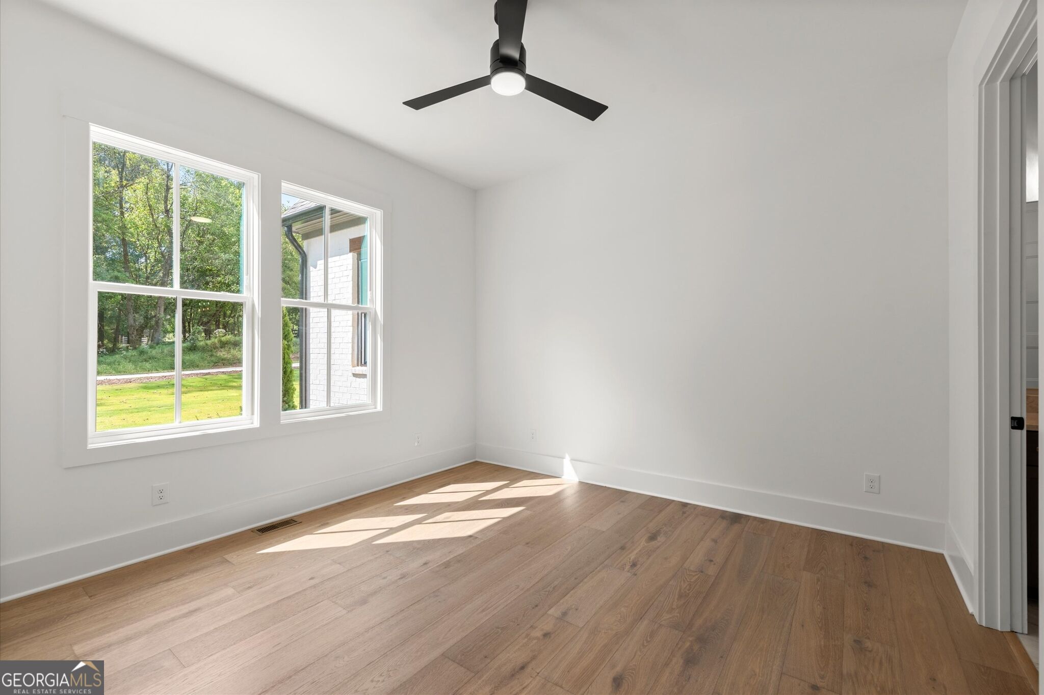 Vacant room with light wood floors, two yard-facing windows, and a black ceiling fan.