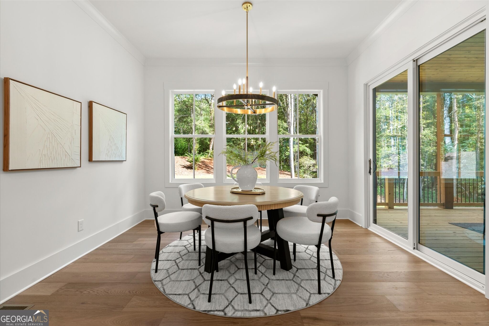 Dining room with a round table, ring chandelier, and windows overlooking woods and a deck.