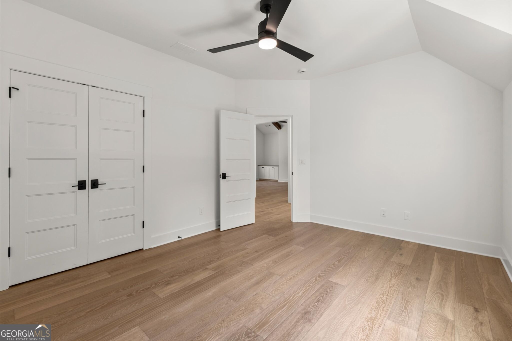 Empty room with vaulted ceiling, wood floors, white double doors, and a black ceiling fan.
