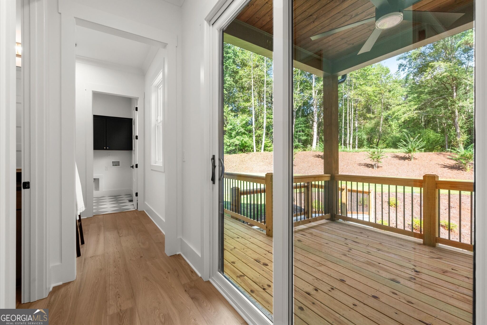 Hallway opening onto a covered wooden deck with a ceiling fan and wooded yard beyond.