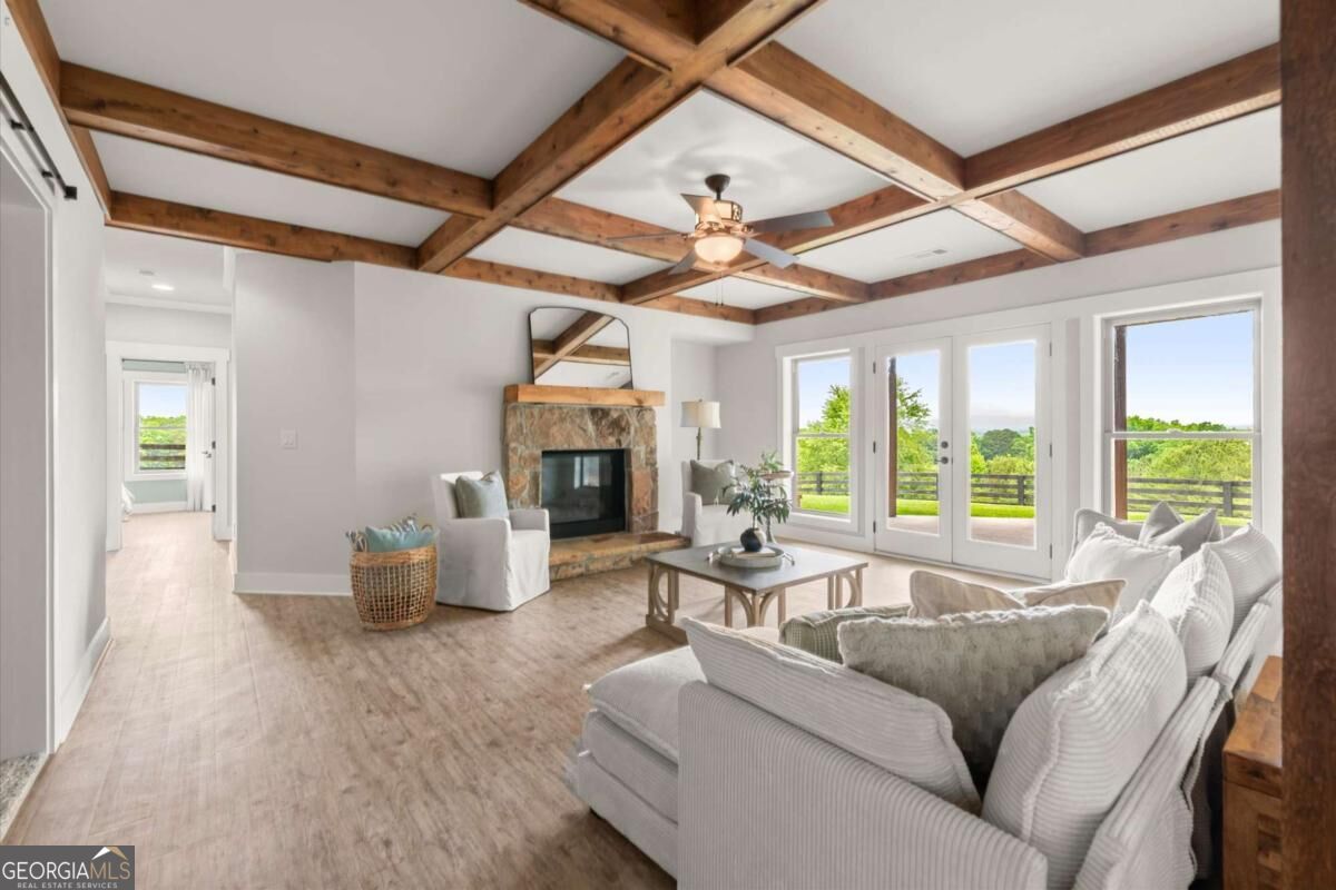 Living room with exposed timber ceiling beams, stone fireplace, and windows overlooking greenery.