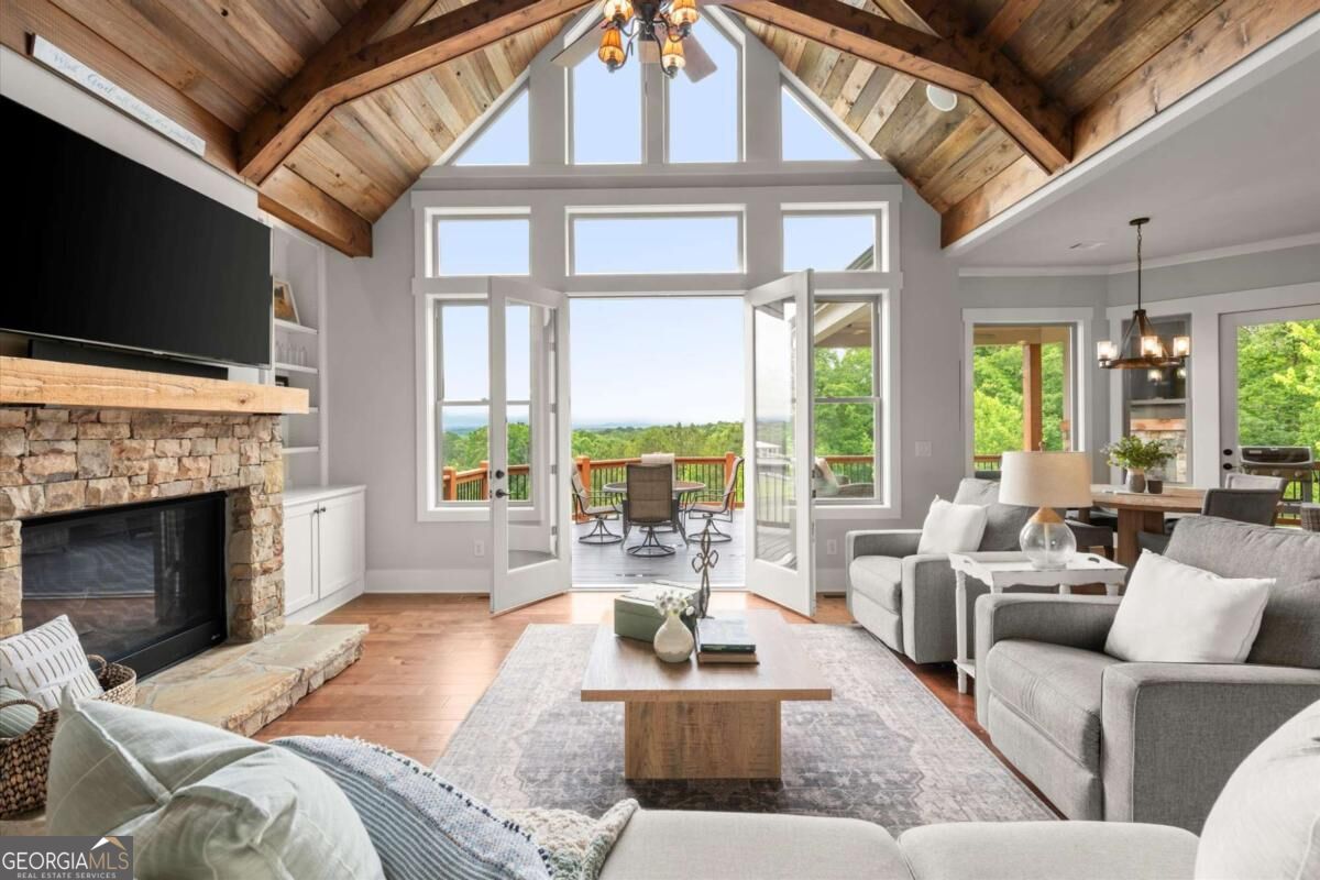 Living room with a vaulted wood ceiling, stone fireplace, and open doors to a wooded-view deck.