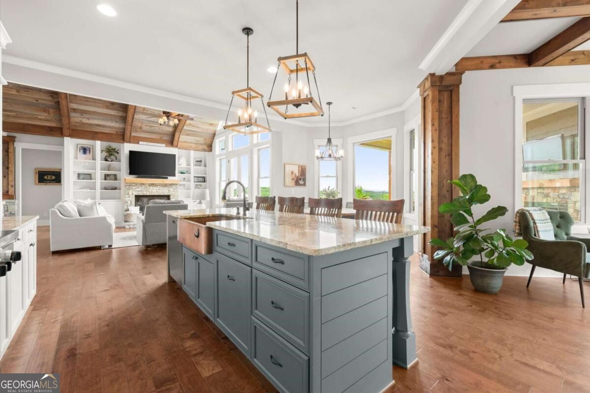 Open kitchen with a large gray island, stone countertop, exposed wood beams, and adjoining living room.