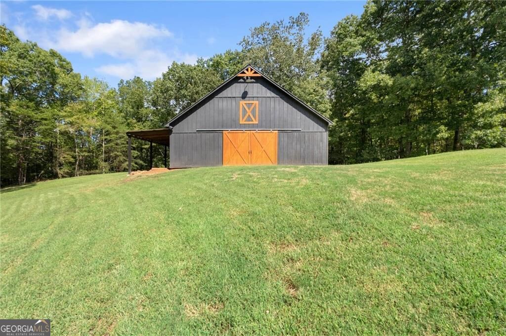 Dark gray barn with orange doors on a sloping lawn, with a covered lean-to and trees behind it.