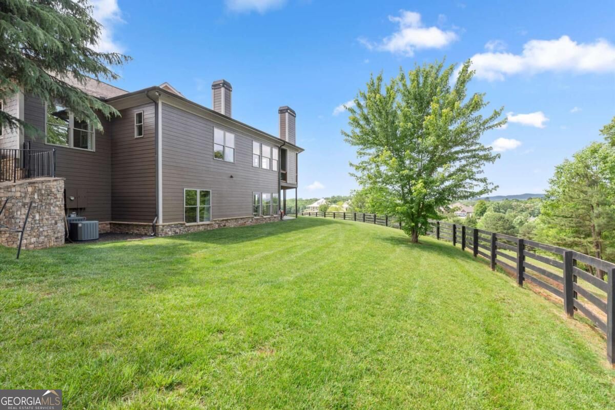Two-story home beside a fenced lawn, with a leafy tree and distant wooded hills.