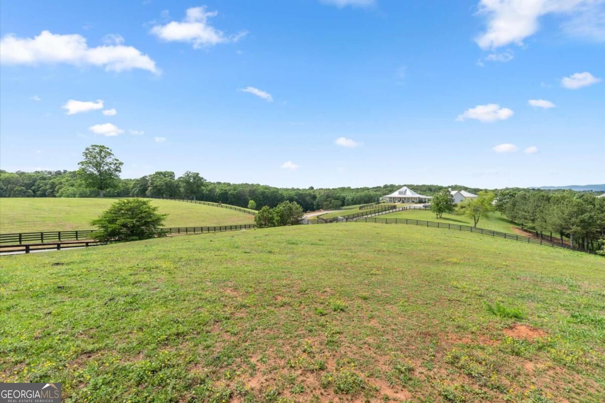 Open grassy acreage with black fencing, scattered trees, and distant buildings.