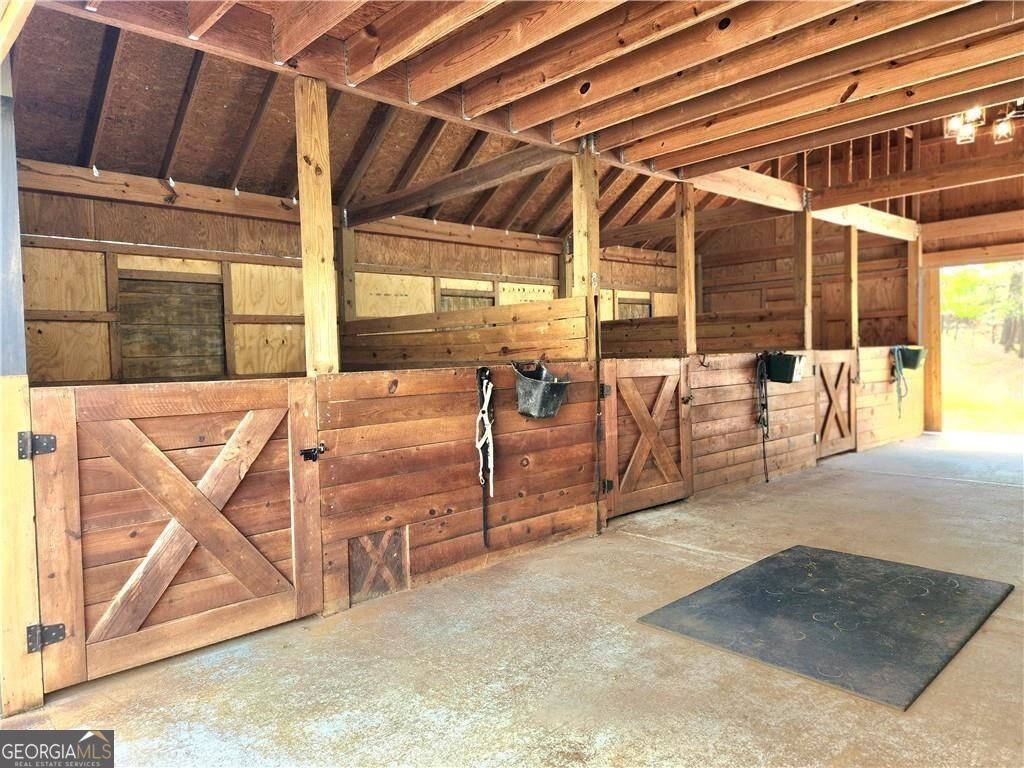 Barn interior with wooden horse stalls, cross-braced doors, exposed rafters, and a concrete floor.
