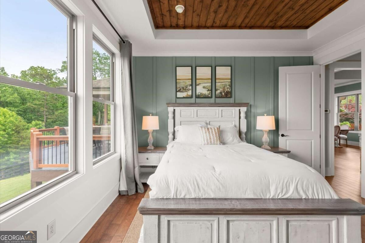 Bedroom with a white bed, green paneled wall, wood ceiling, and large windows overlooking trees and a deck.