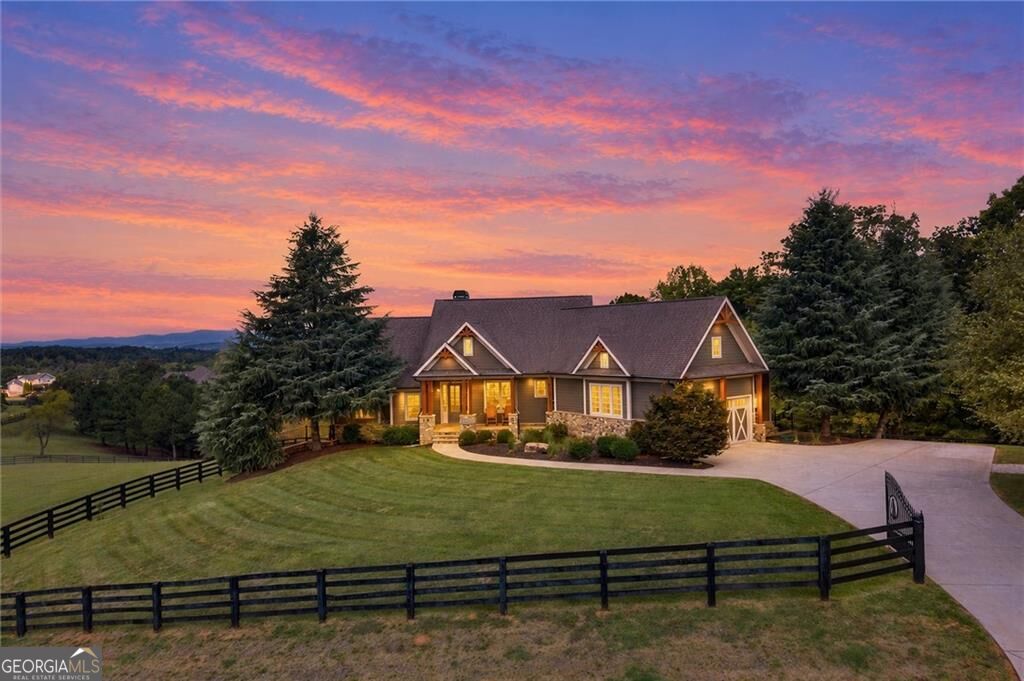 Gray gabled house behind a fenced lawn, framed by evergreen trees under a vivid sunset sky.