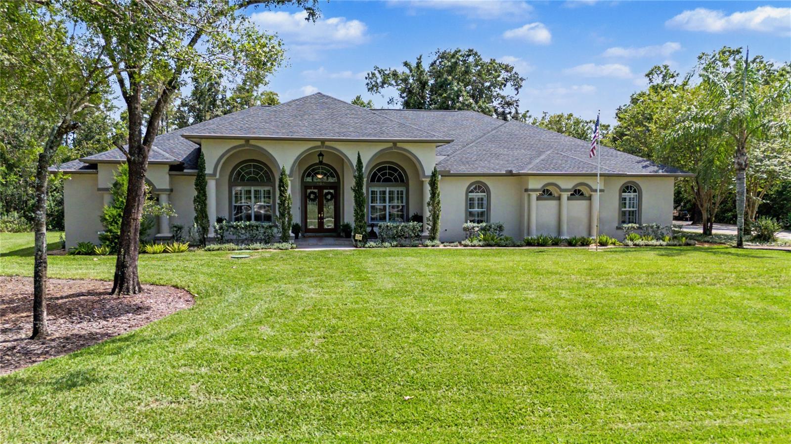 Cream stucco home with three arched entry bays, framed by mature trees and a broad lawn.